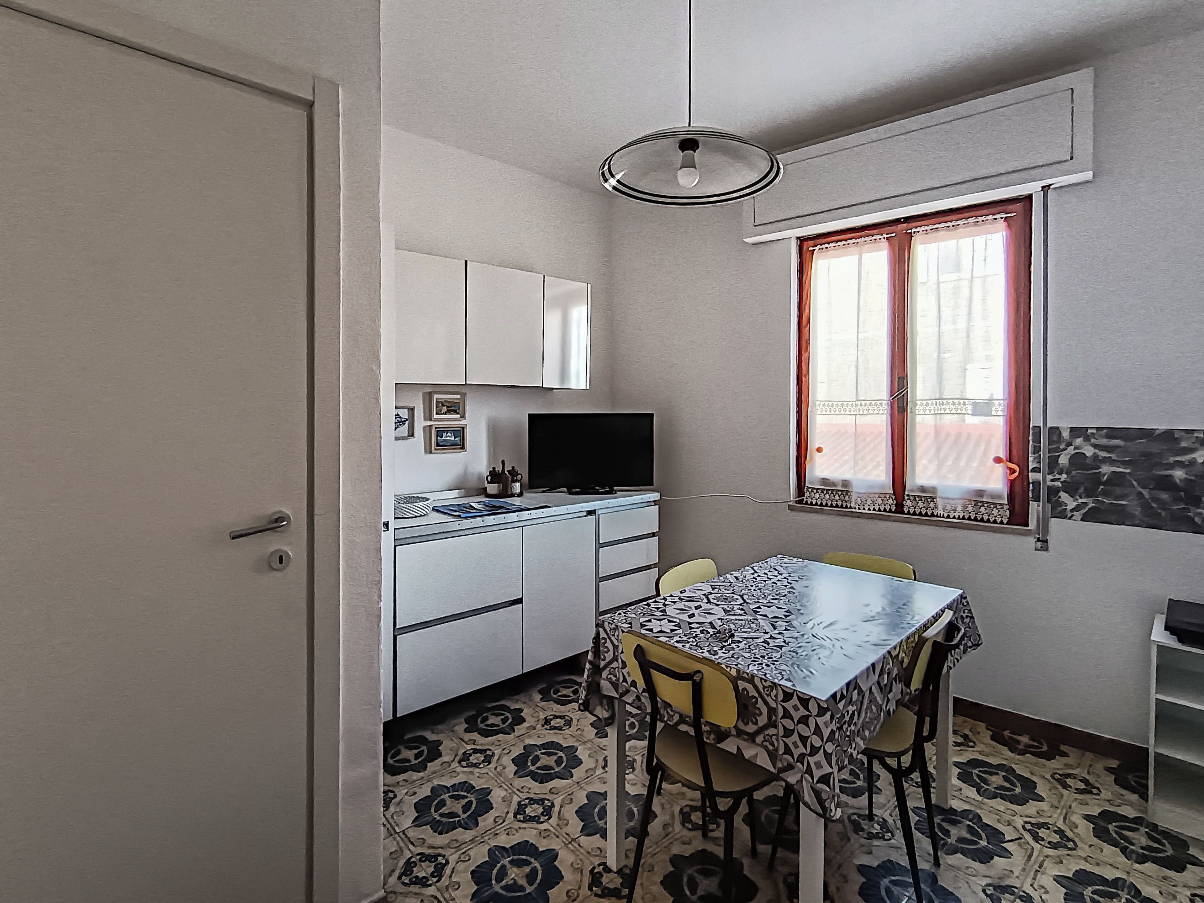 A compact kitchen with white cabinets, a dining table with chairs, a television, and a window with curtains.