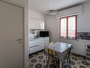 A compact kitchen with white cabinets, a dining table with chairs, a television, and a window with curtains. - Casa Lara (Carloforte)