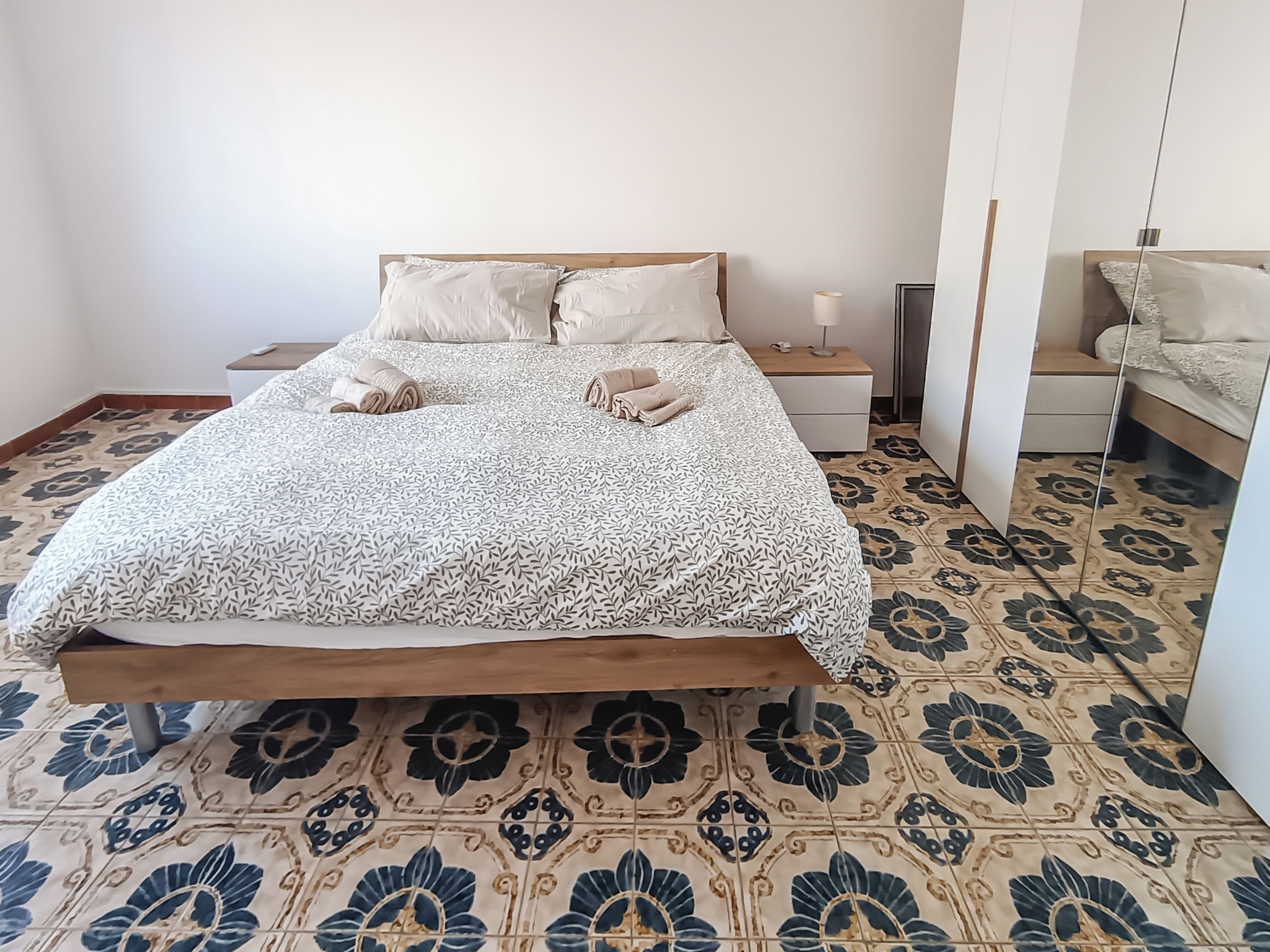 Uma cama de solteiro com colcha estampada, mesa de cabeceira com abajur e um espelho.