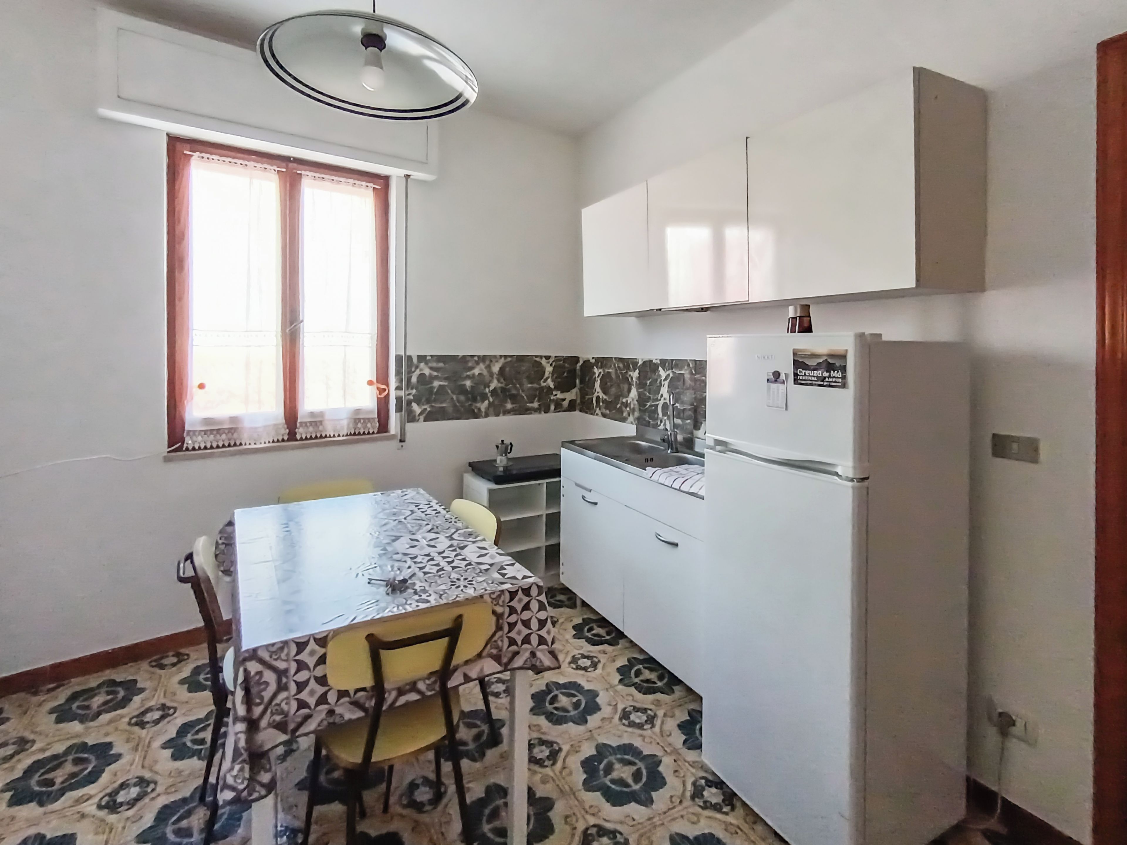 A compact kitchen with a dining table, chairs, a refrigerator, and a window with curtains.