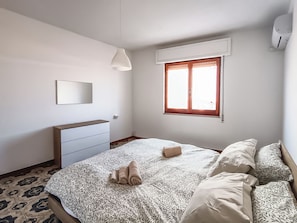 A bedroom with a bed, a bedside table, a mirror, and a window with blinds. - Casa Lara (Carloforte)