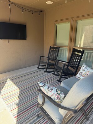 Ein sonniger Patio-Bereich mit an der Wand befestigtem Fernseher, zwei Schaukelstühlen und einem gemusterten Teppich.