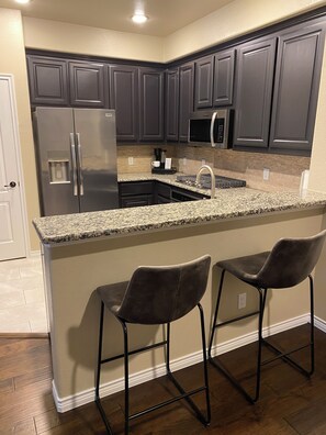A kitchen with dark cabinets, a stainless steel refrigerator, a microwave, and a granite countertop. There are two bar stools with metal frames. - 3-bedroom townhome in Plano’s entertainment and corporate area. Comfy Texas vibe (Plano)