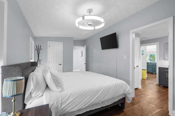 A bedroom with a bed, a bedside table, a lamp, a flat-screen TV, and a door leading to another room.
