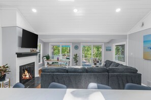 A modern living room with a fireplace, a large gray sectional sofa, a flat-screen TV mounted above the fireplace, and a dining area with a table and chairs.