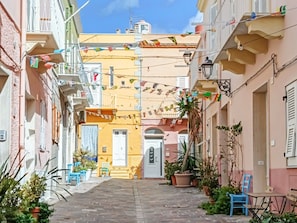 A narrow street with buildings on either side, a central door, and potted plants. - Casa Lara - Charming Apartment in the Heart of Carloforte's Historic Center (Carloforte)