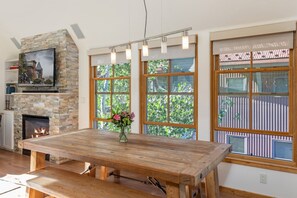 A spacious room with a large wooden table, a stone fireplace, and large windows with a view of greenery.