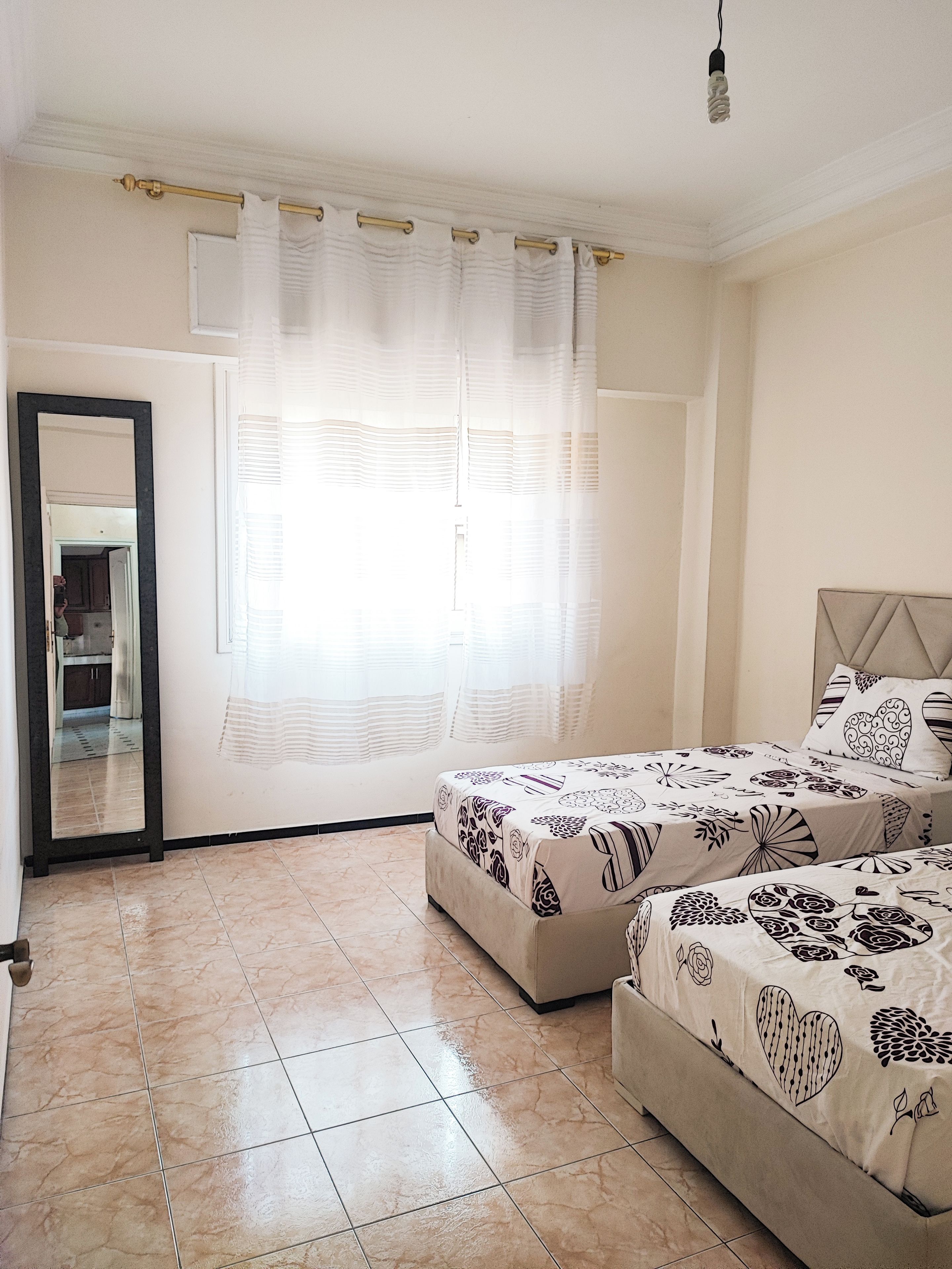 A bedroom with two beds, a large mirror, and a window with sheer curtains.