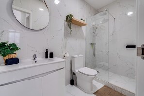 Une salle de bain avec un lavabo, des toilettes et une douche, le tout en marbre blanc. On y trouve un miroir, une étagère avec une plante et un panier contenant une plante.