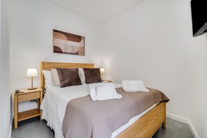 A neatly made bed with a wooden headboard, two bedside tables with lamps, a wall-mounted TV, and a painting above the bed. - Casa Sol e Sal  3-Min Walk to Beach & Old Town (Albufeira)