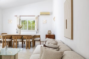 A modern living room with a glass coffee table, a beige sofa, a dining area with wooden chairs, and a window with curtains. - New! Quinta Lamy - Villa with Pool near Carvoeiro (Lagoa)