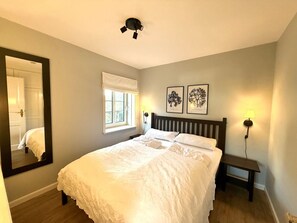 A bedroom with a bed, a nightstand, a wall-mounted lamp, a window with a white valance, and two framed wall art pieces. - Deichhof - Apt. 18 (Dunsum)
