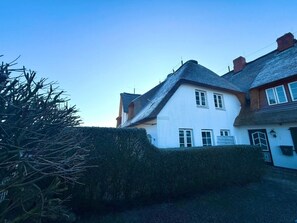 A white house with a steeply pitched roof, a hedge in the foreground, and a clear sky. - Deichhof - Apt. 18 (Dunsum)