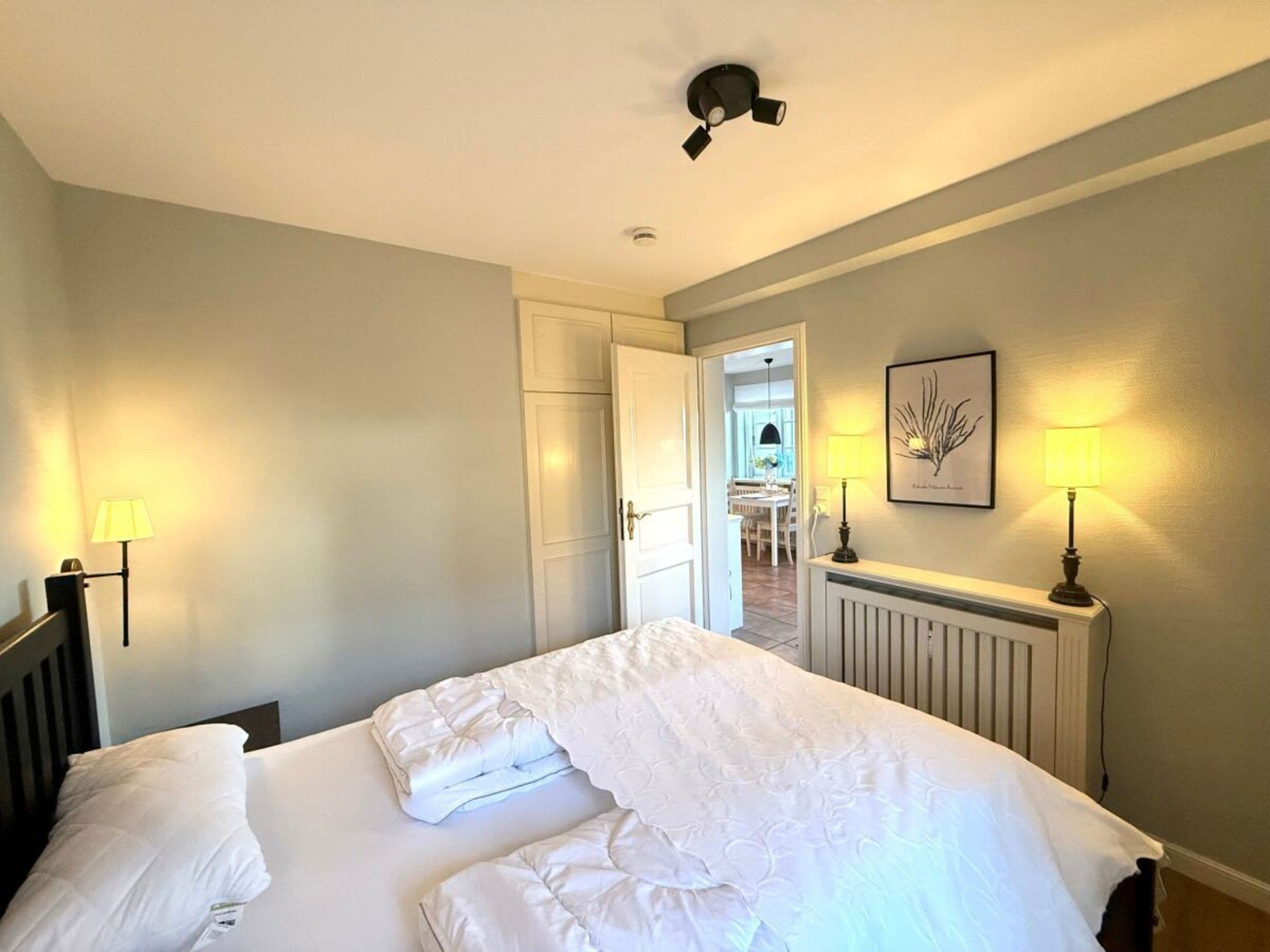Quarto de hotel com cama, abajures, ventoinha no teto e porta que dá para uma sala de jantar.