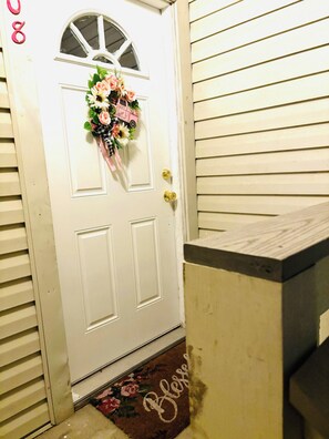 Une porte blanche avec une fenêtre, un paillasson orné de motifs floraux et une petite voiture rose en guise de décoration.