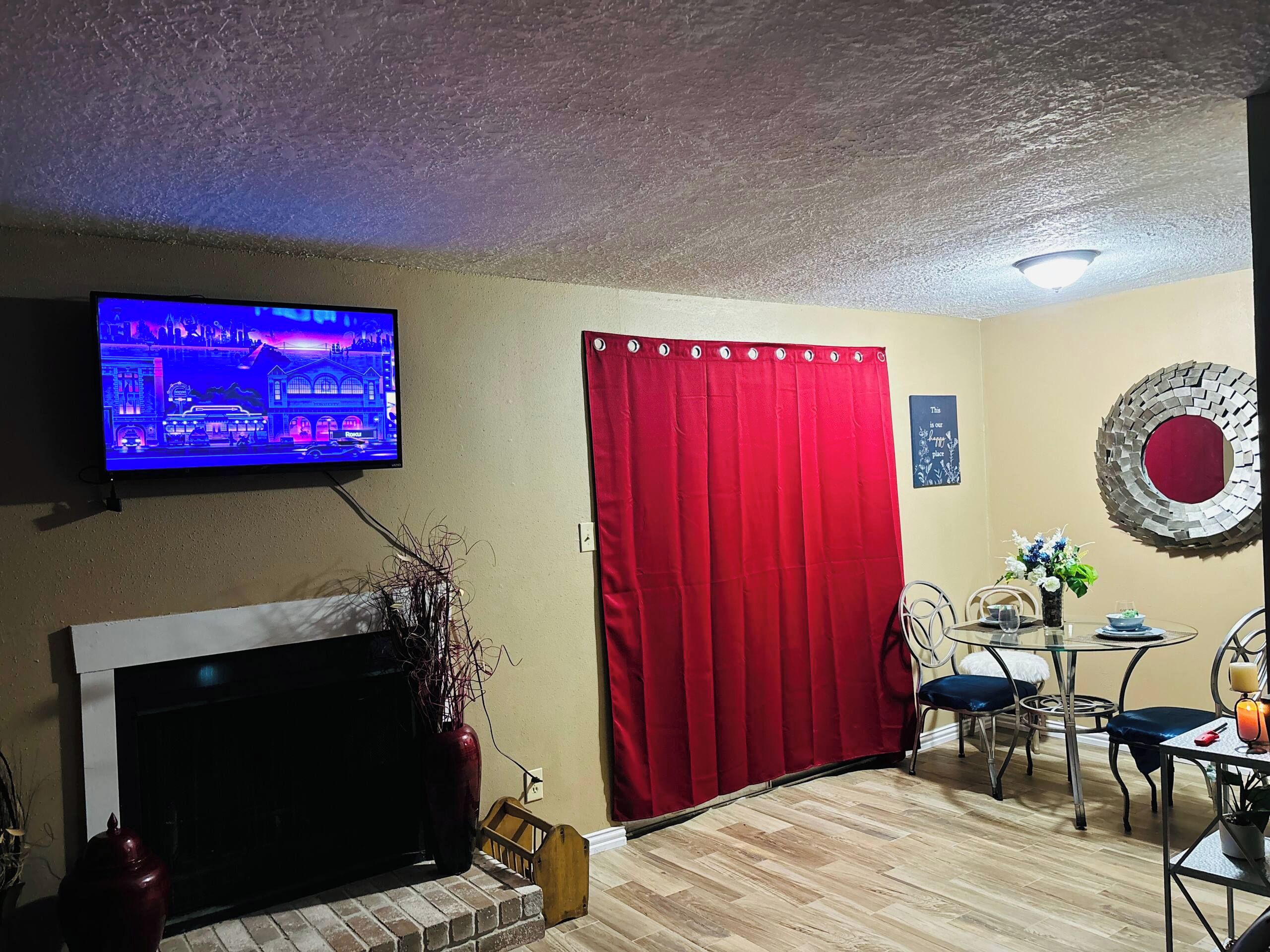 Una sala de estar con televisión, chimenea, mesa de comedor con sillas, cortina roja y espejo decorativo.
