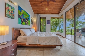 A bedroom with a large bed, a ceiling fan, and a view of the outdoors.