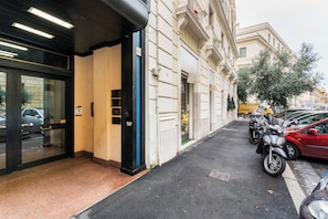 Una entrada de edificio con un puerta de vidrio, una puerta negra y una puerta azul. Hay coches aparcados y una furgoneta amarilla en la calle.