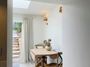 A room with a table and chairs, a window with curtains, a wall-mounted light fixture, and a view of a garden outside. - Redwood Cottage (Bristol)