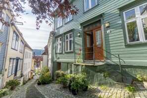 Un carrer pavimentat i estret amb edificis de tipus tradicional europeu, una porta de fusta i una petita zona de jardí amb plantes en testos.