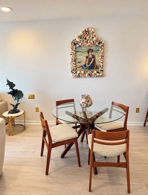 A dining area with a glass table surrounded by wooden chairs, a decorative painting with a seashell border, and a small side table with a plant. - Upscale Oceanfront Penthouse Villa Sleeps 4 Panoramic Ocean Views. (Sawgrass)