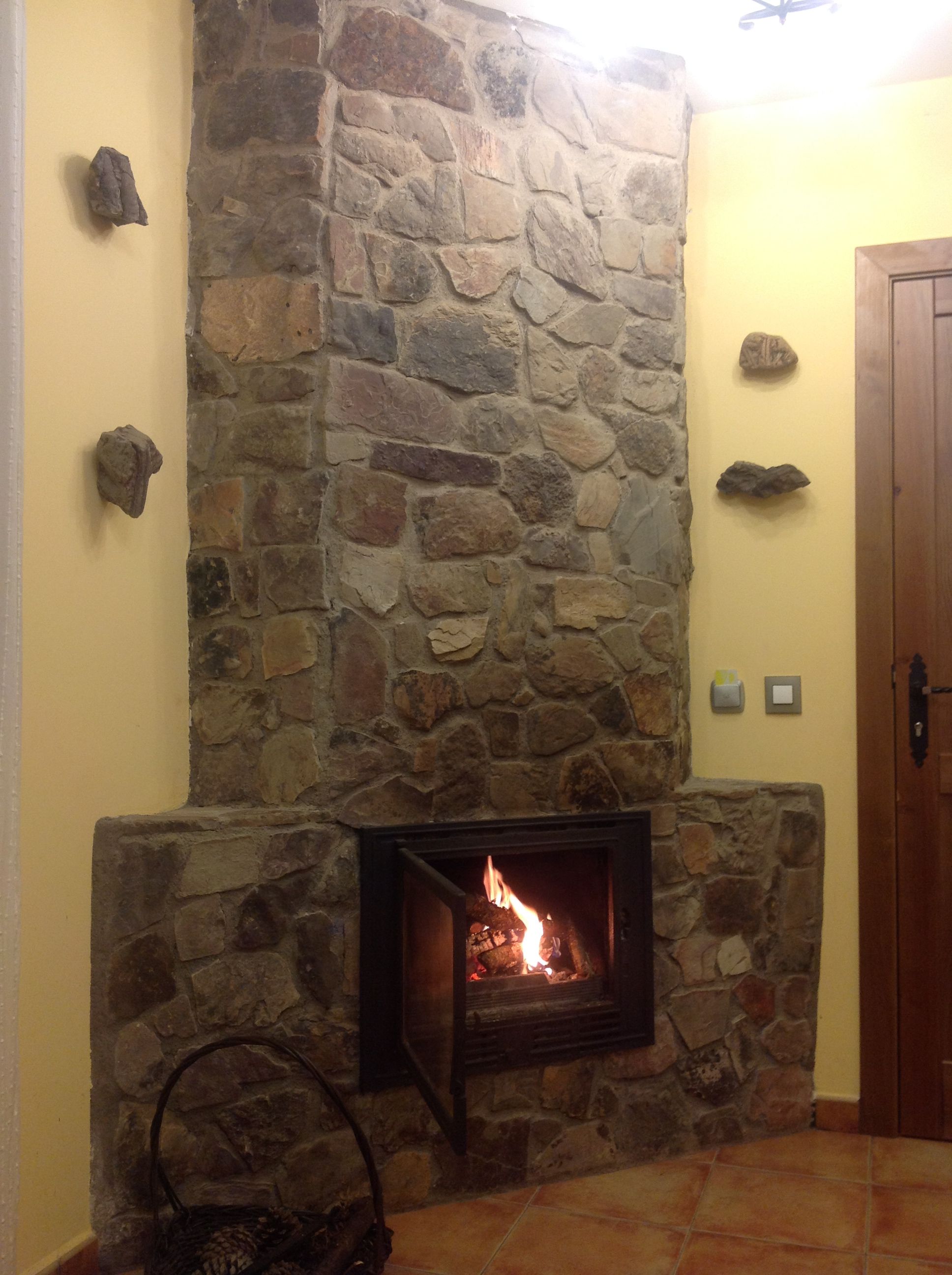 A stone fireplace with a fire, a door to the right, and a stone wall with rocks on it.