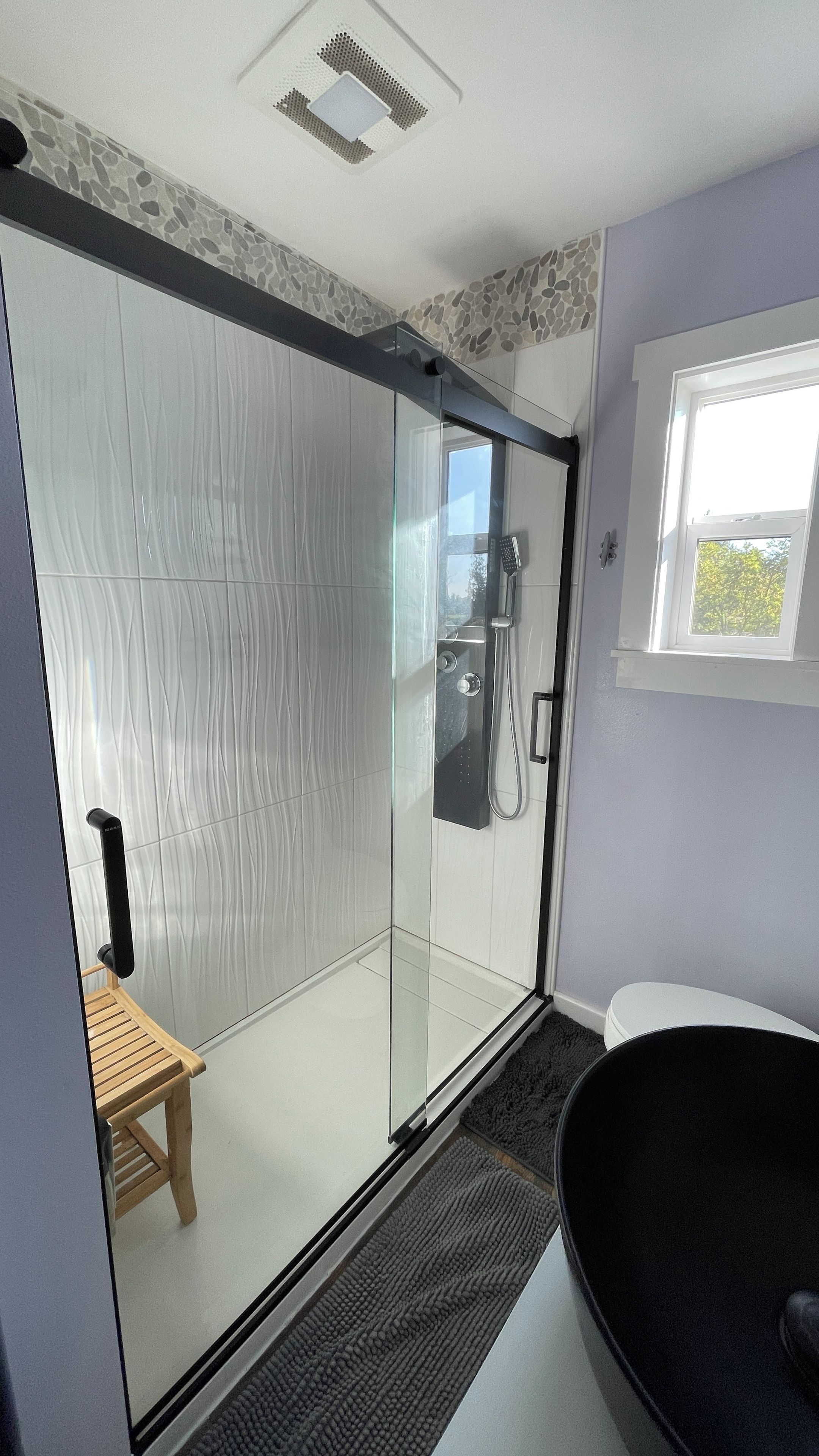 A modern bathroom with a glass shower enclosure, a wooden bench, a bathtub, and a window providing natural light.