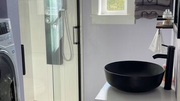 A modern kitchen with a glass shower door, a black bowl on the countertop, and a window with a view of greenery.