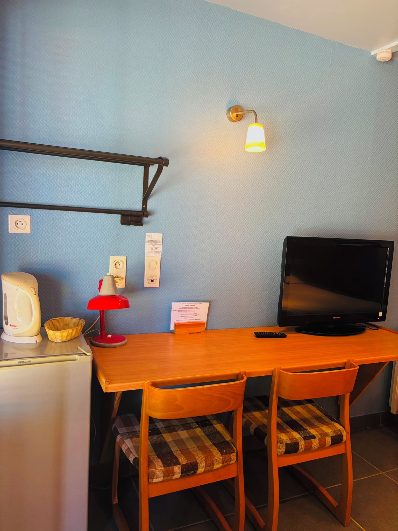 Habitació senzilla amb escriptori de fusta, dues cadires, televisió, làmpada vermella, una paperera i una làmpada penjant a la paret.