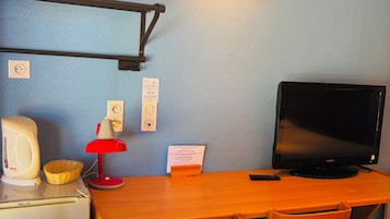 Habitació senzilla amb escriptori de fusta, dues cadires, televisió, làmpada vermella, una paperera i una làmpada penjant a la paret.