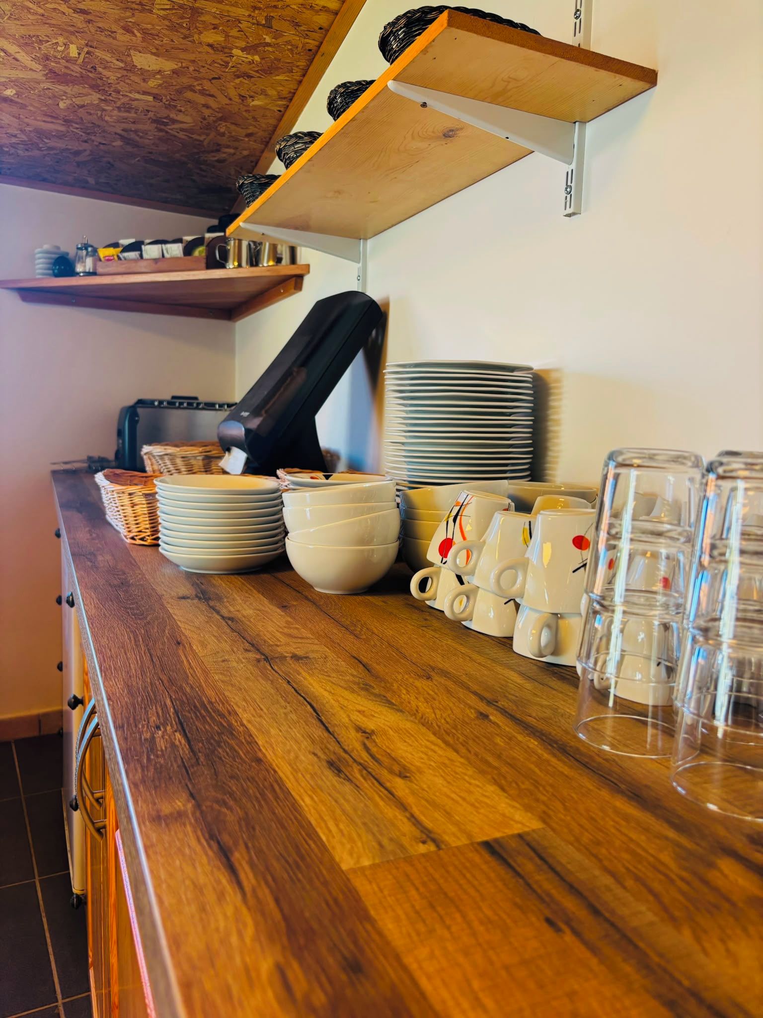 Una encimera de cocina con platos, tazones y tazas apilados, una máquina de café y un estante de madera arriba.