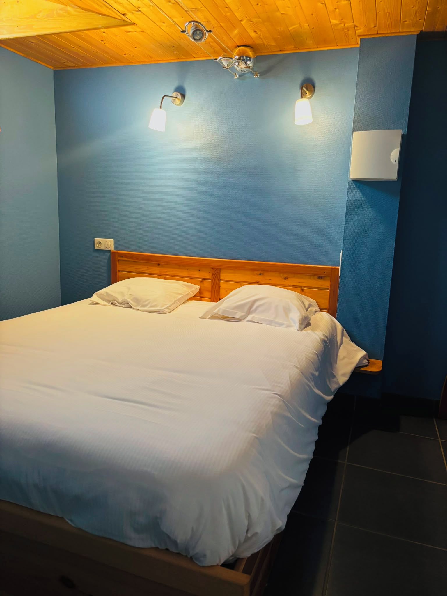 Una habitación con cama de cabecera de madera, dos lámparas fijadas a la pared y un termostato.