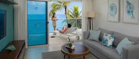 A living room with a view of the ocean, a balcony with a person sunbathing, and a sofa with decorative pillows.