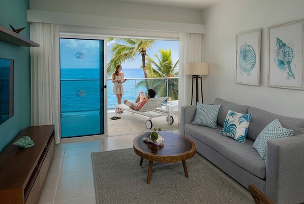 Una sala de estar con vista al océano, un balcón con una persona tomando el sol y un sofá con almohadones decorativos.