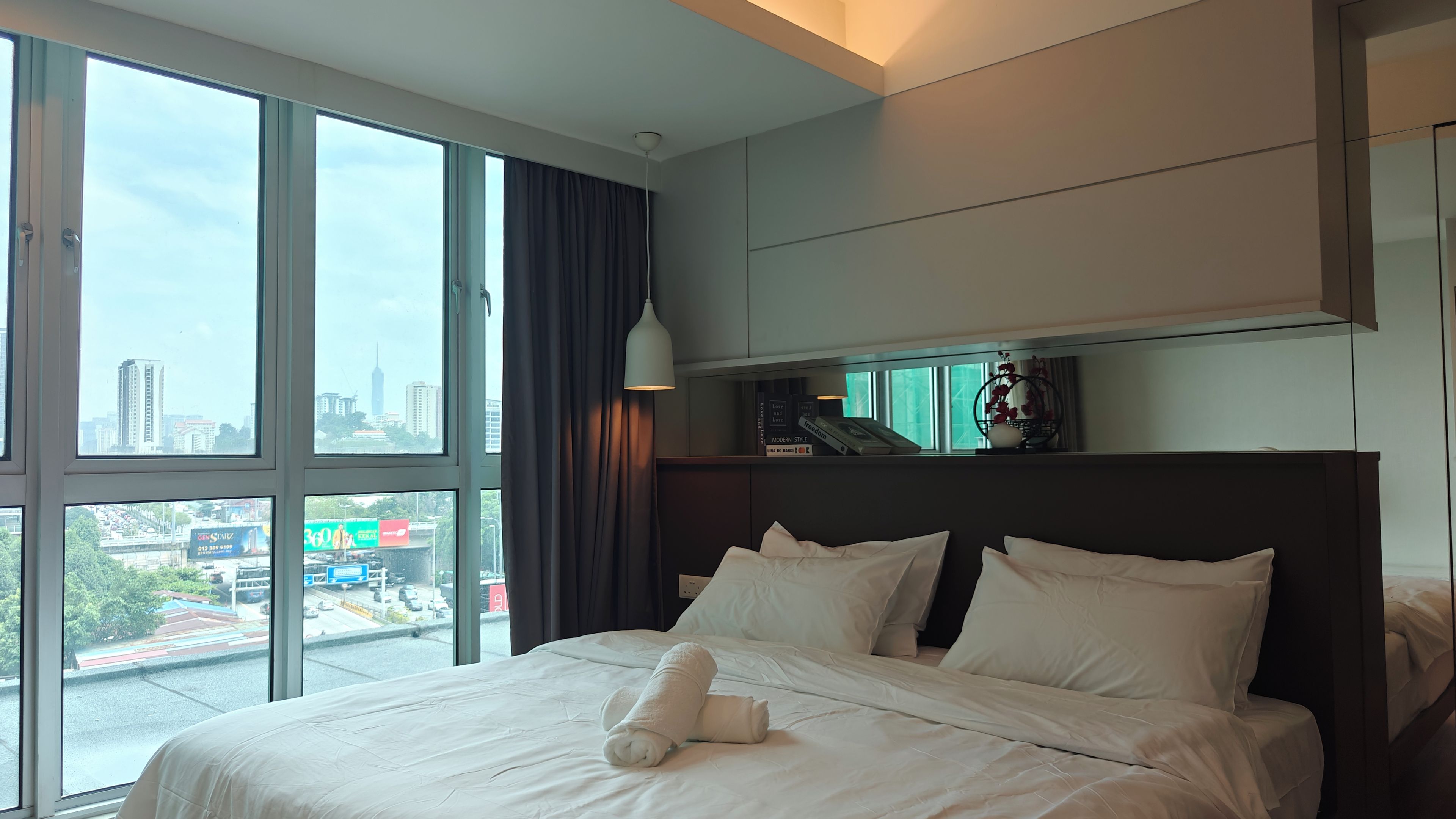 Ein Hotelzimmer mit einem großen Bett, einem Nachttisch, einer Lampe, einem Fenster mit Stadtblick und einem Wandregal mit Büchern und Dekoration.