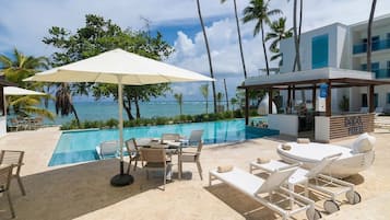 A poolside area with lounge chairs, a table, and umbrellas, overlooking a pool and the ocean.