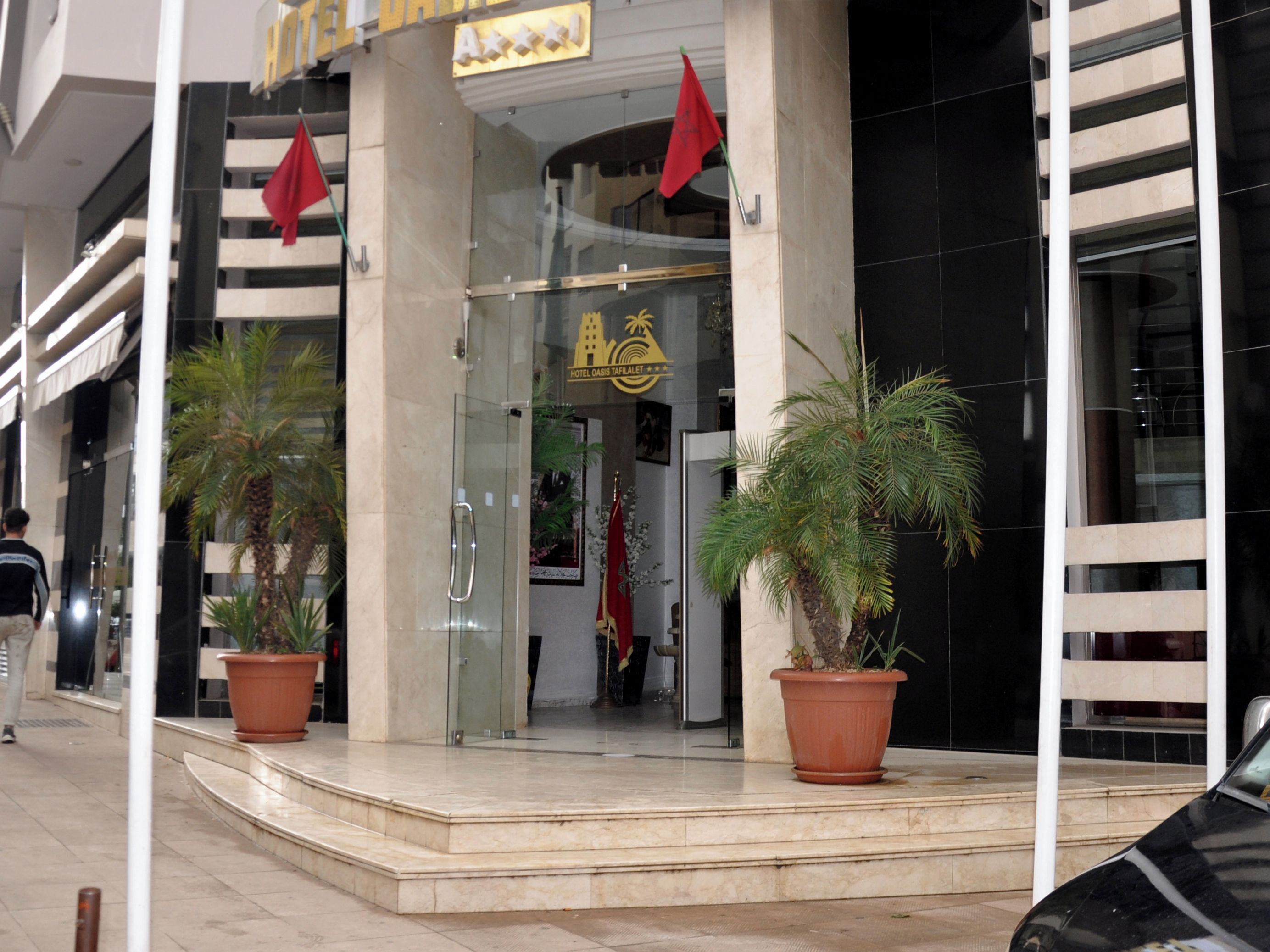 A hotel entrance with a glass door, palm trees, and red flags.