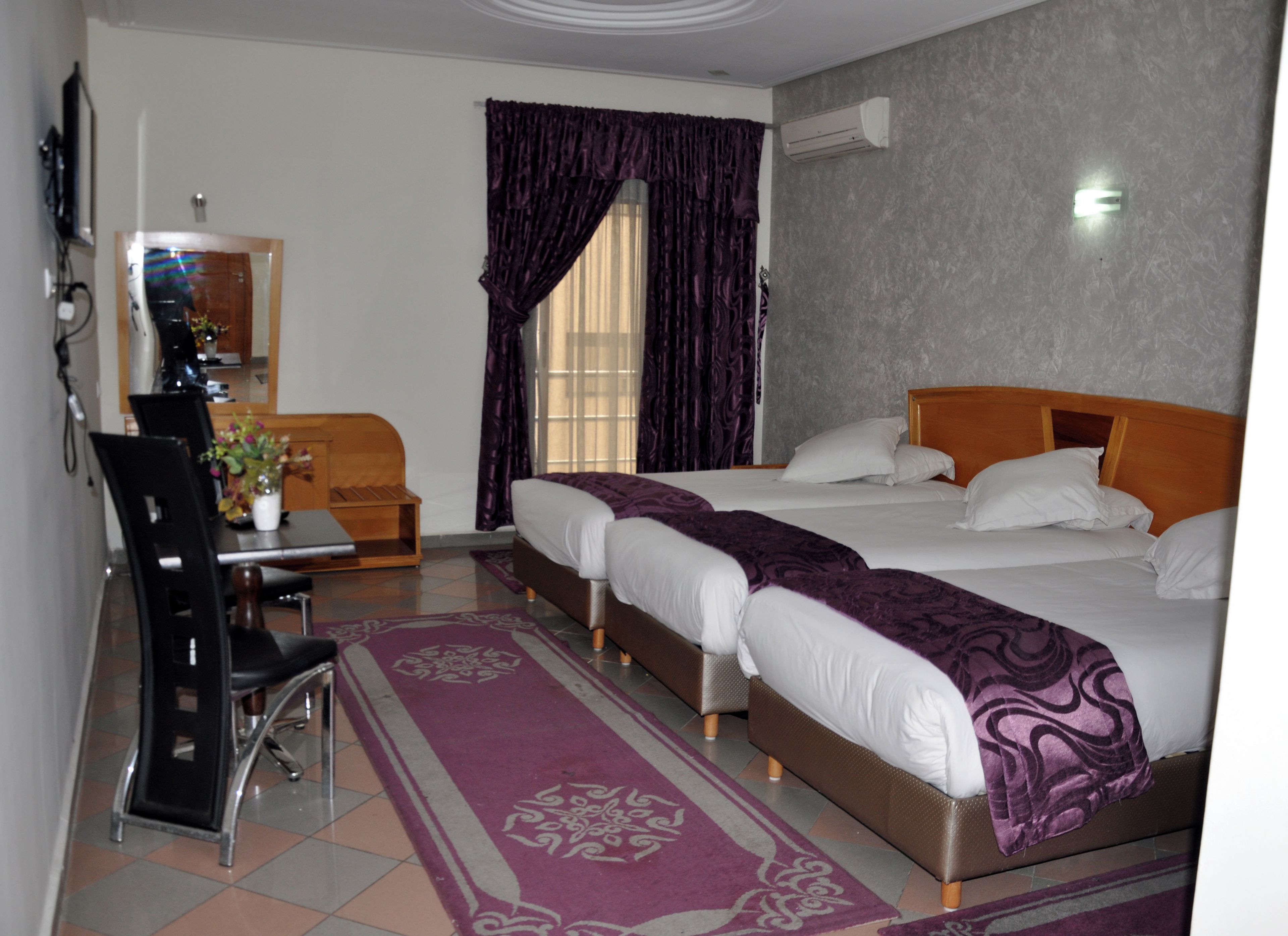 Une chambre d’hôtel comprenant un lit, deux tables de chevet, un bureau avec une chaise, une fenêtre avec des rideaux et un appareil de climatisation.