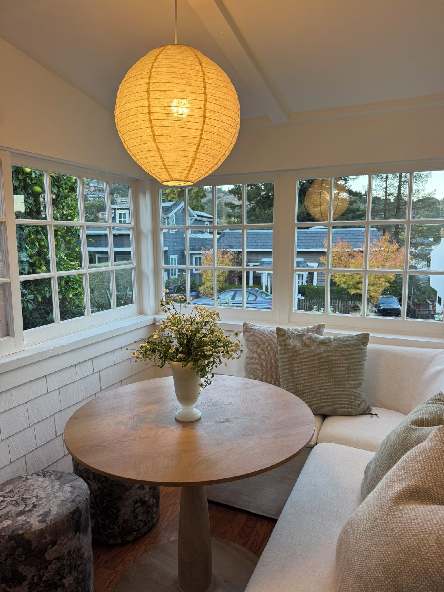 A room with a round wooden table, a white vase with flowers, a white sofa, and a large window with a view of houses and trees.