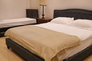 Ein Doppelbett mit dunklem Kopfteil und einer beigen Decke, ein Nachttisch mit Lampe sowie ein kleines Regal.