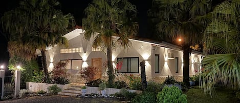 A two-story house with a well-lit exterior, surrounded by palm trees and a gravel driveway.