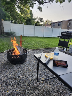 A backyard with a fire pit, a grill, and a table with a bottle and a phone. - Charming 3-bedroom apartment in lovely Utica (Utica)