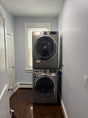 A laundry room with two stacked front-loading washing machines, a window with blinds, and a door. - Charming 3-bedroom apartment in lovely Utica (Utica)