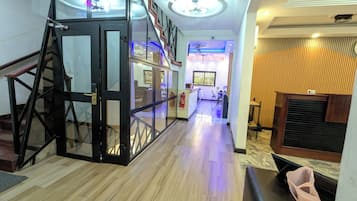 Vestíbulo de hotel con ascensor de vidrio, una recepción y un extintor.