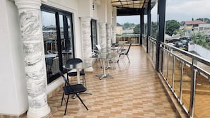 A balcony with a tiled floor, metal-framed tables and chairs, and large windows with a view of the city.