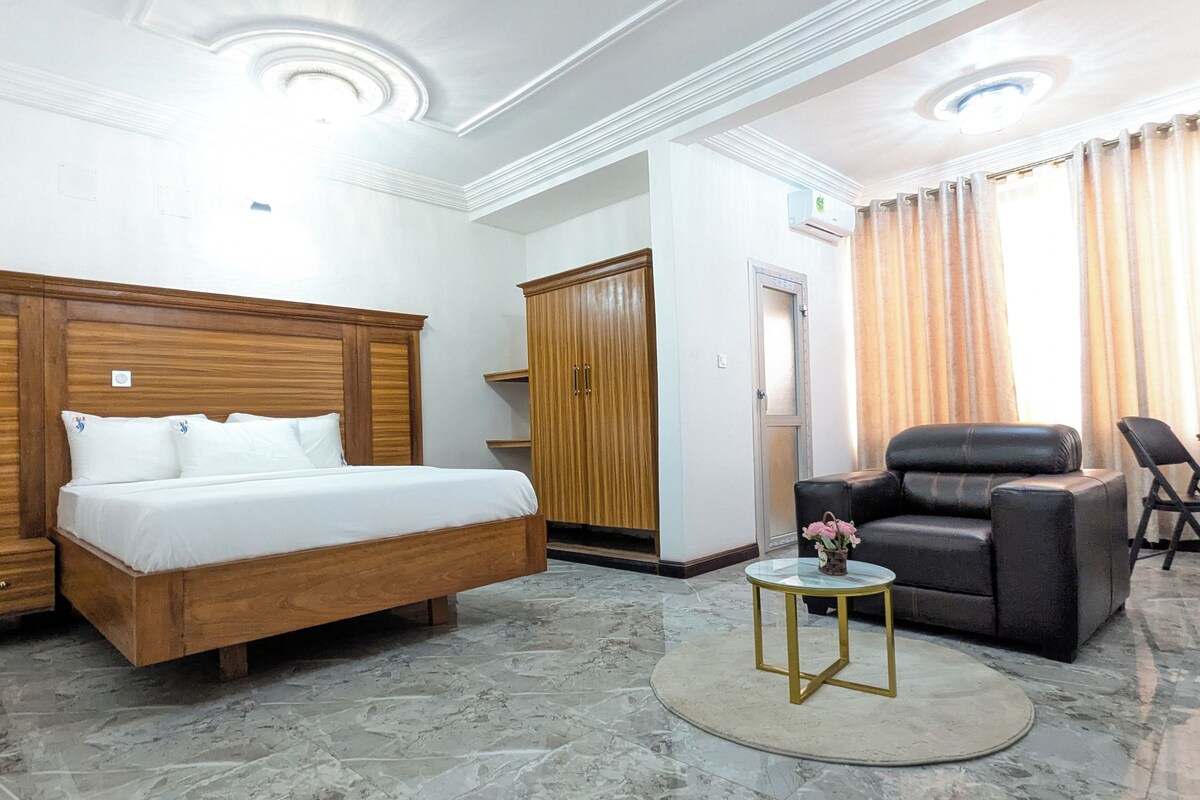 Een hotelkamer met een bed, een zithoek met een stoel en tafel, een kledingkast en een bijzettafeltje met een vaas.
