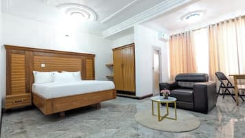 Habitación de hotel con cama, zona de estar con una silla y mesa, armario y una mesita de noche con un florero.
