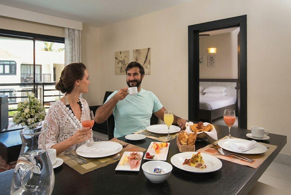 A hotel room with a dining table set for breakfast, including plates of food, a pitcher, and a glass of orange juice.