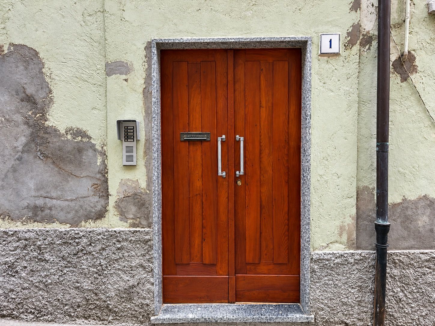 Una puerta doble de madera con un picaporte metálico y una placa, ubicada en una pared con textura y un cartel con un número.