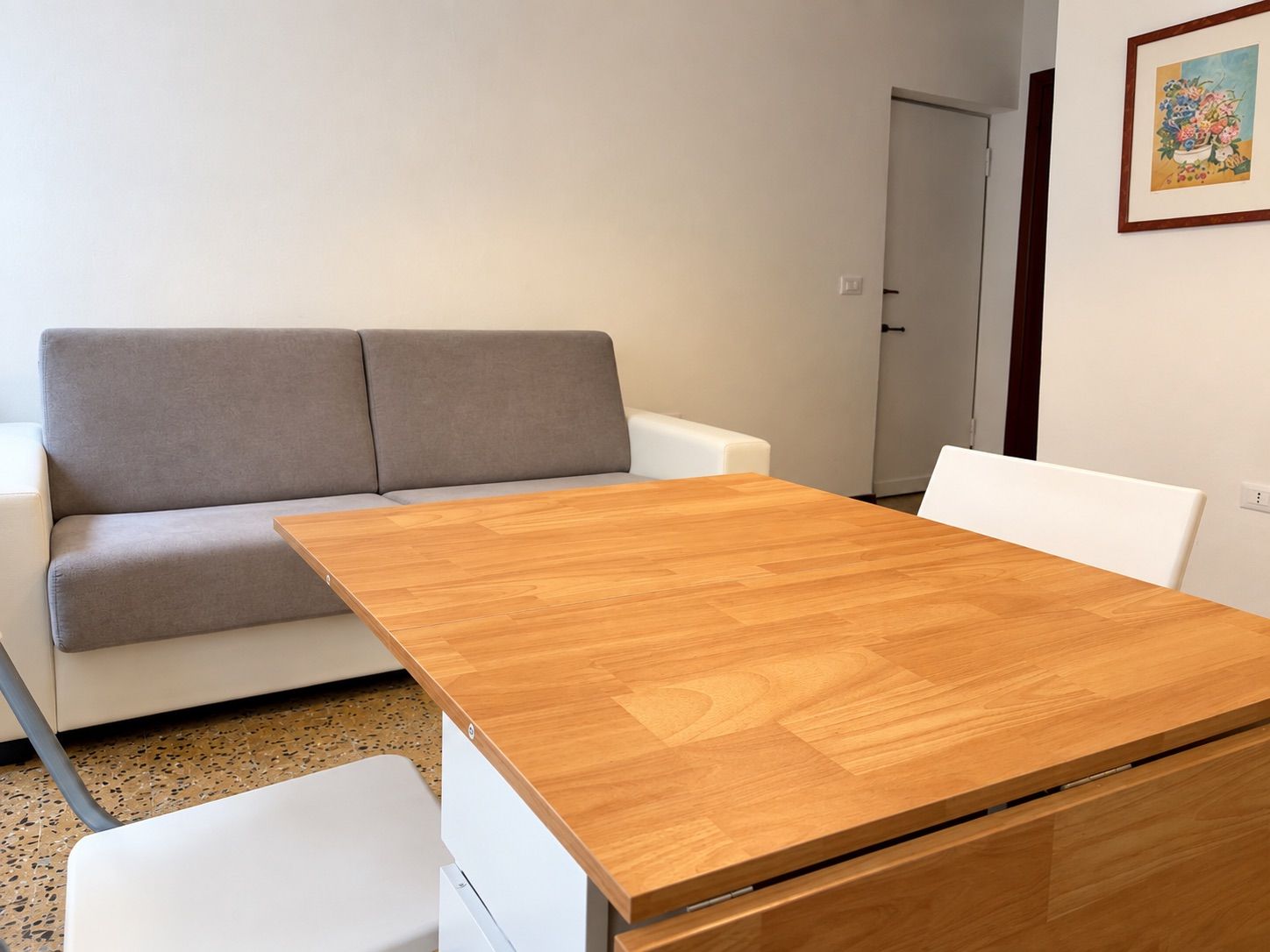 A room with a wooden table, white chairs, a grey sofa, and a door.